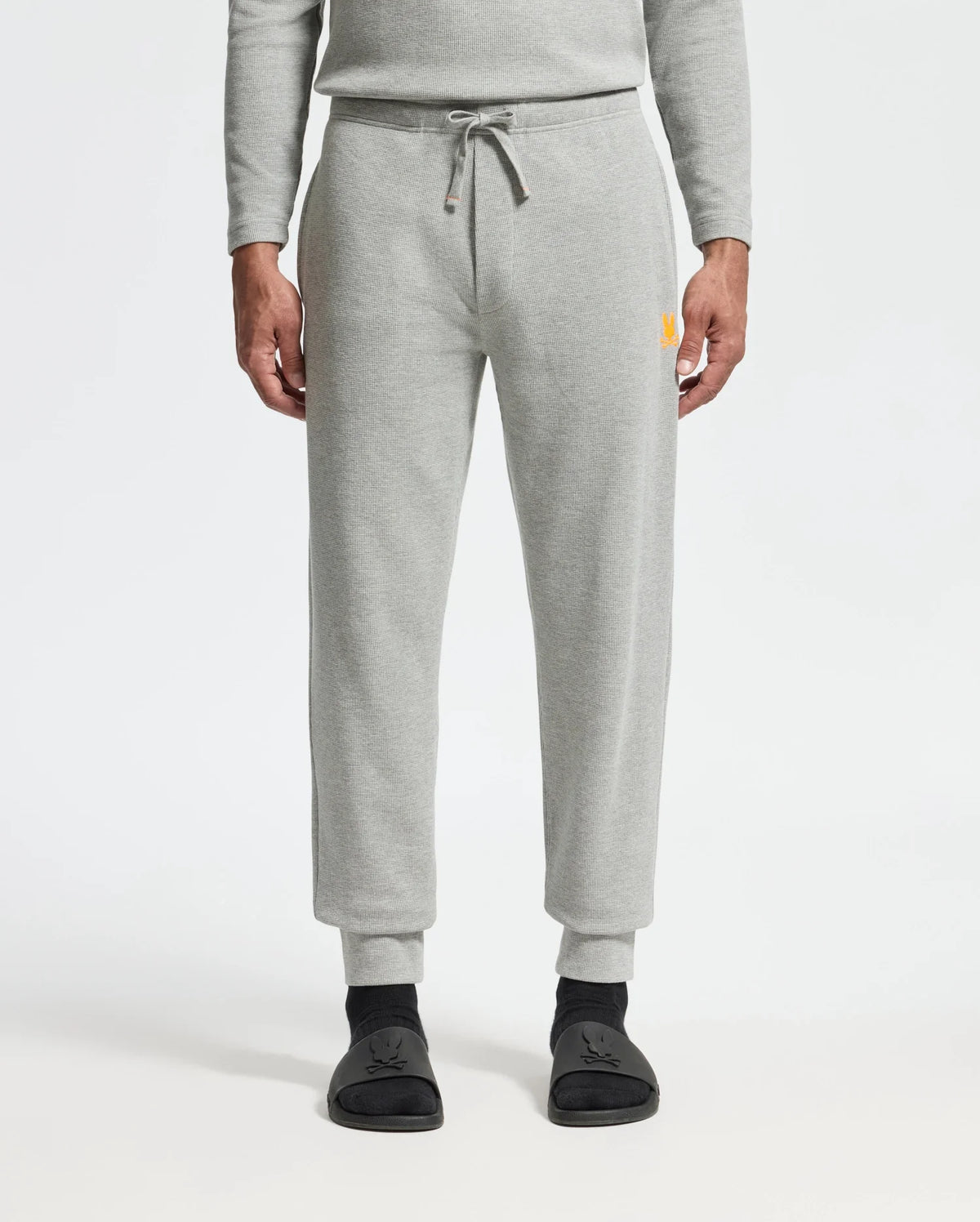 A person wearing the light gray Psycho Bunny MENS VAL WAFFLE LOUNGE PANT - B6P673E200 and a matching top stands with hands at their sides. The pants feature a drawstring waist and have a small yellow logo on the upper right leg. Black socks with a subtle design are visible, all set against a plain white background that enhances the outfit's simplicity.
