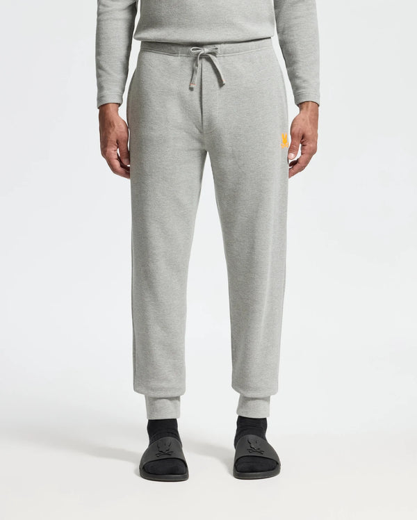 A person wearing the light gray Psycho Bunny MENS VAL WAFFLE LOUNGE PANT - B6P673E200 and a matching top stands with hands at their sides. The pants feature a drawstring waist and have a small yellow logo on the upper right leg. Black socks with a subtle design are visible, all set against a plain white background that enhances the outfit's simplicity.