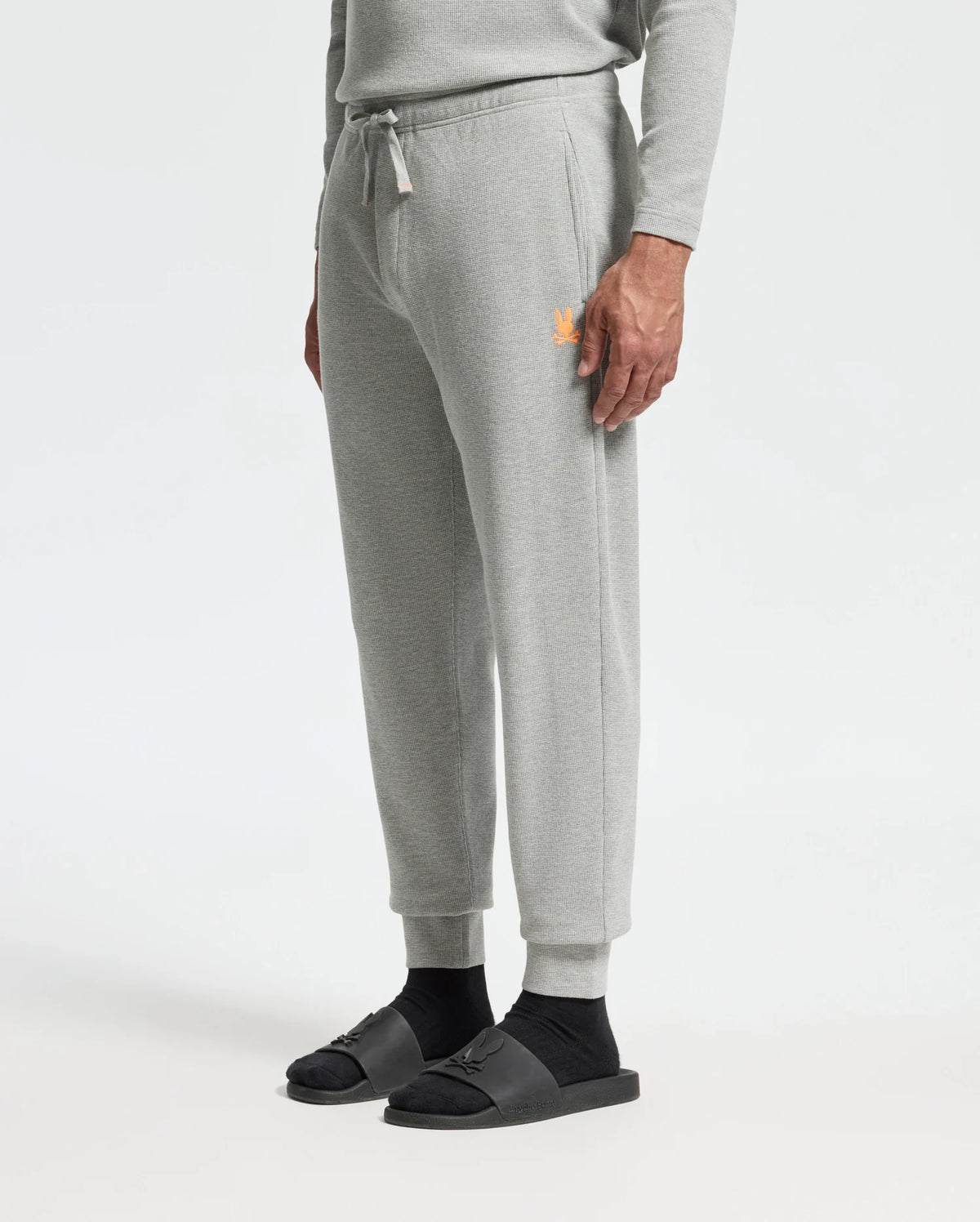 A person is dressed in the MENS VAL WAFFLE LOUNGE PANT by Psycho Bunny, which features a matching light gray waffle knit sweatshirt. The pants are adorned with an orange logo on the left side. They complete their outfit with black socks and dark gray slide sandals against a plain white background.