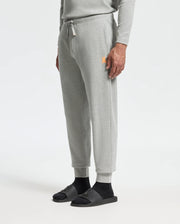A person is dressed in the MENS VAL WAFFLE LOUNGE PANT by Psycho Bunny, which features a matching light gray waffle knit sweatshirt. The pants are adorned with an orange logo on the left side. They complete their outfit with black socks and dark gray slide sandals against a plain white background.