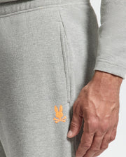 A close-up features someone wearing the MENS VAL WAFFLE LOUNGE PANT - B6P673E200 by Psycho Bunny, showcasing their hand resting by their side. The pants are light gray waffle-knit with an embroidered orange bunny logo near the pocket, seamlessly blending comfort and style.