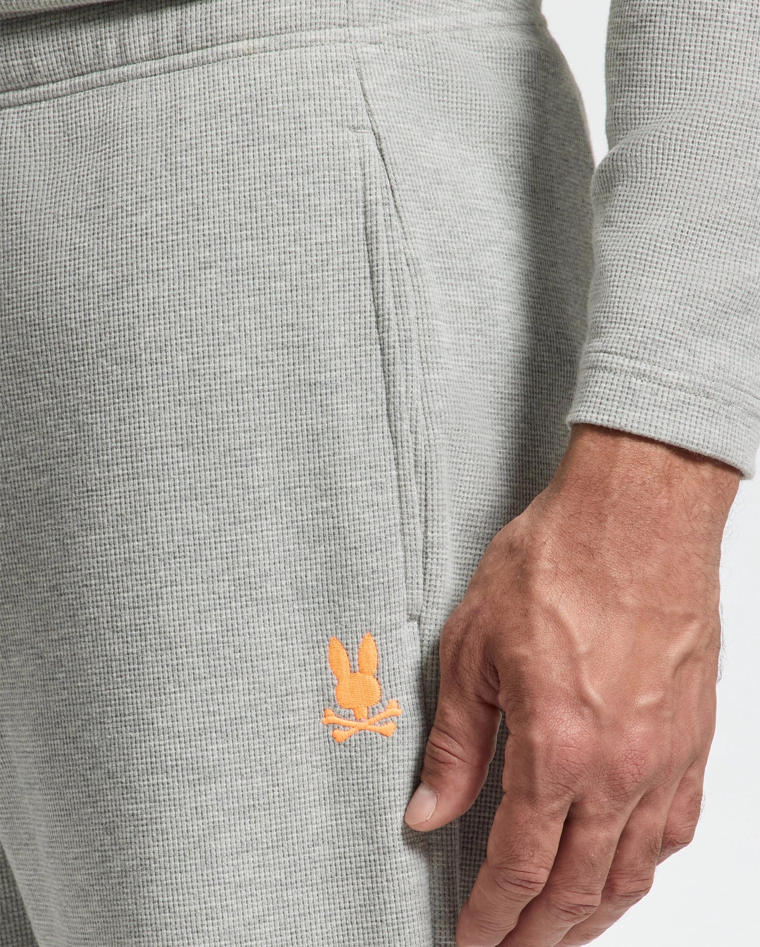 A close-up features someone wearing the MENS VAL WAFFLE LOUNGE PANT - B6P673E200 by Psycho Bunny, showcasing their hand resting by their side. The pants are light gray waffle-knit with an embroidered orange bunny logo near the pocket, seamlessly blending comfort and style.