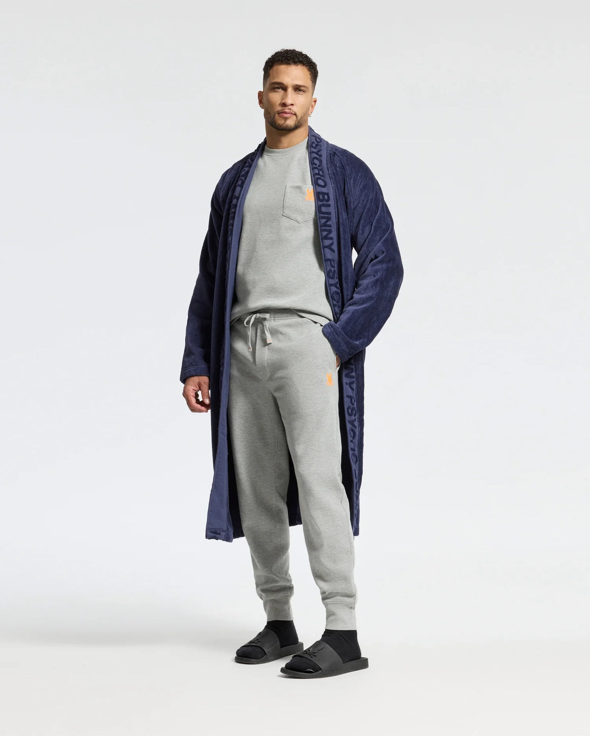 A man exudes relaxed style in a light gray tracksuit and black slides, confidently complemented by a long navy robe with a patterned trim. The plain white background accentuates his graceful demeanor, akin to donning the ultimate comfort of Psycho Bunny's MENS VAL WAFFLE LOUNGE PANT - B6P673E200.
