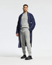 A man exudes relaxed style in a light gray tracksuit and black slides, confidently complemented by a long navy robe with a patterned trim. The plain white background accentuates his graceful demeanor, akin to donning the ultimate comfort of Psycho Bunny's MENS VAL WAFFLE LOUNGE PANT - B6P673E200.
