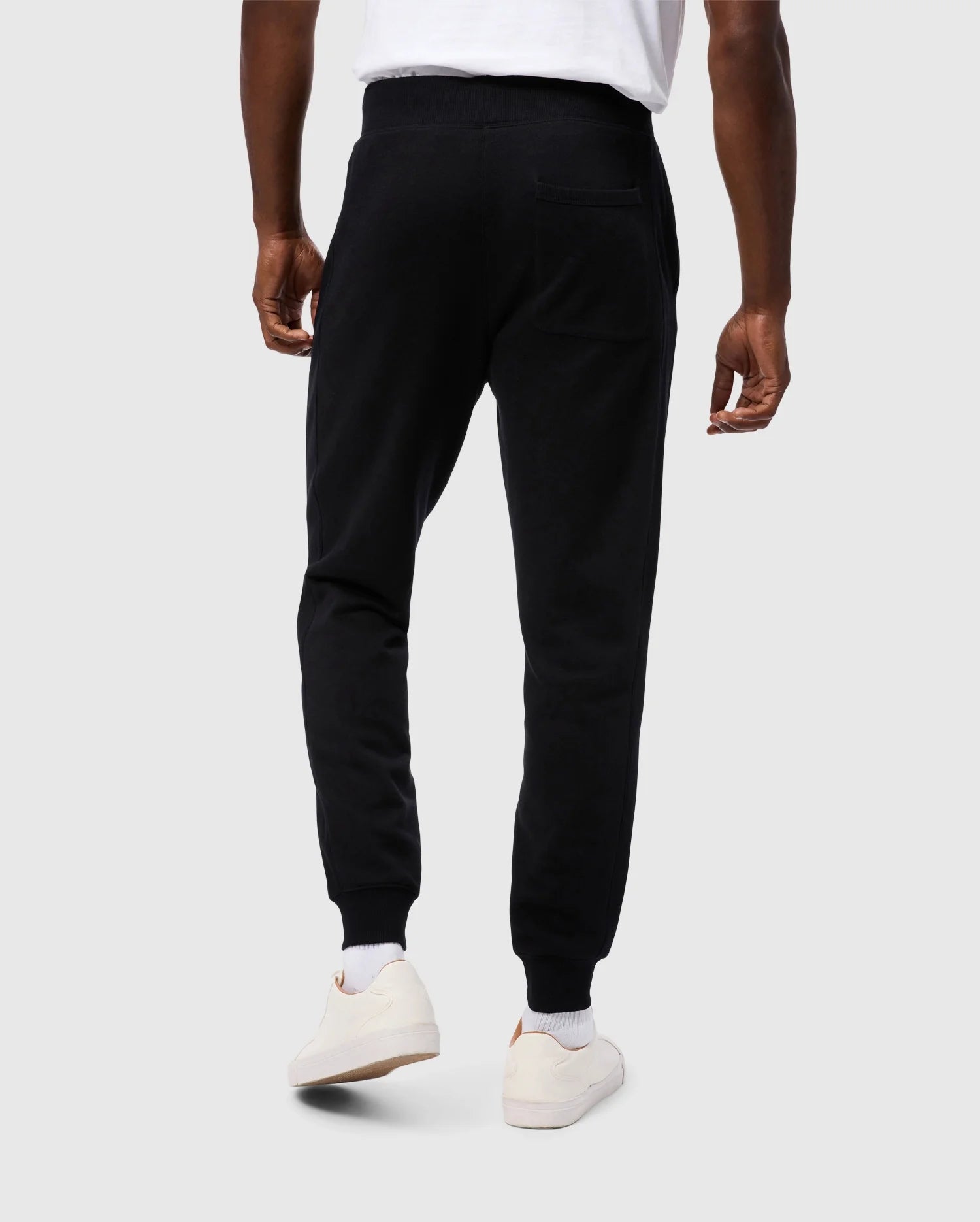 A person is standing facing away from the camera, wearing Psycho Bunny MENS LEVY ESSENTIAL FLEECE SWEATPANT in black (B6P721D200), white socks, and white sneakers. They are also wearing a white t-shirt. The background is a plain, light grey color.