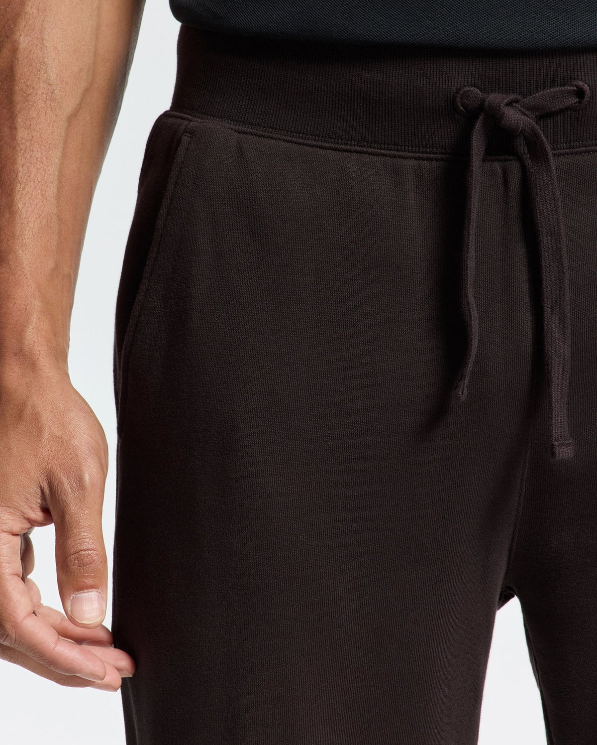 A close-up shows a person wearing Psycho Bunny's Mens Levy Essential Fleece Sweatpant - B6P721D200. Their right hand rests on the side pocket, highlighting the casualwear style against a plain background.