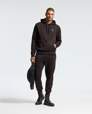 A person in a black hoodie and Psycho Bunny's MENS LEVY ESSENTIAL FLEECE SWEATPANT (B6P721D200) holds a black bag. They wear black boots, standing against a plain light background.