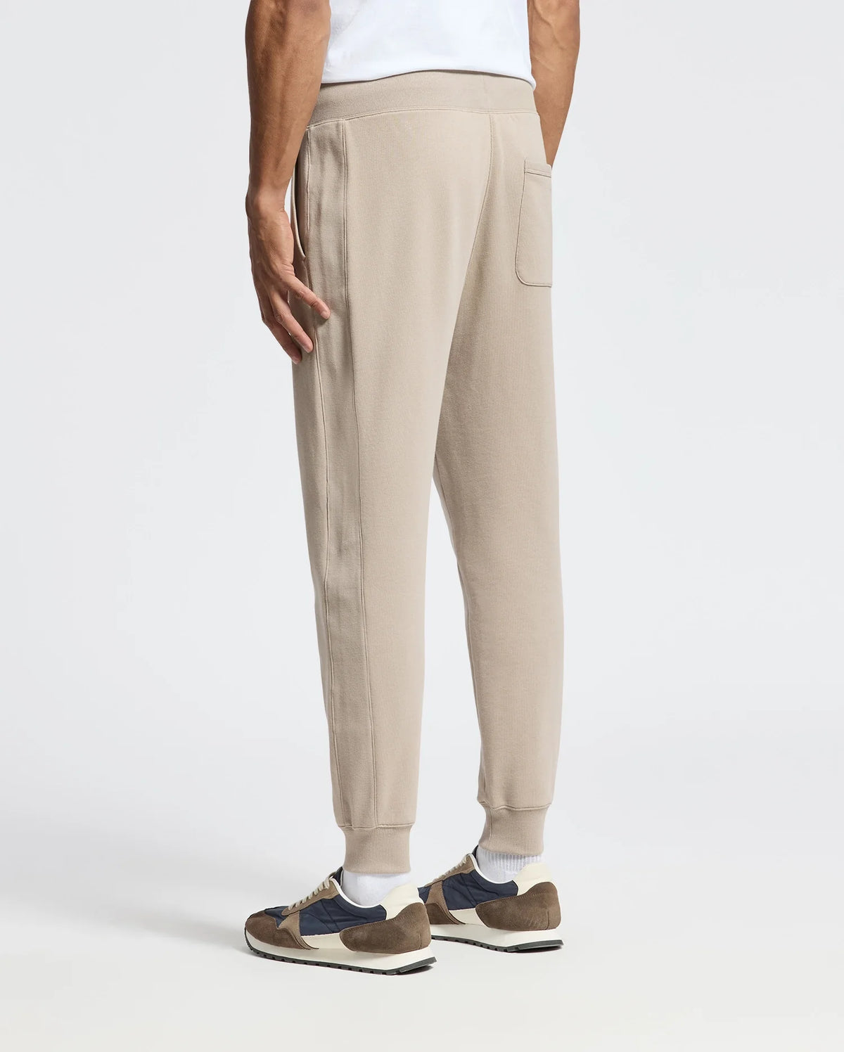 A person dressed in the Psycho Bunny MENS LEVY ESSENTIAL FLEECE SWEATPANT - B6P721D200, paired with a white shirt and multicolored sneakers, stands on a plain white background. The view from behind emphasizes the relaxed fit and single back pocket of the sweatpants.