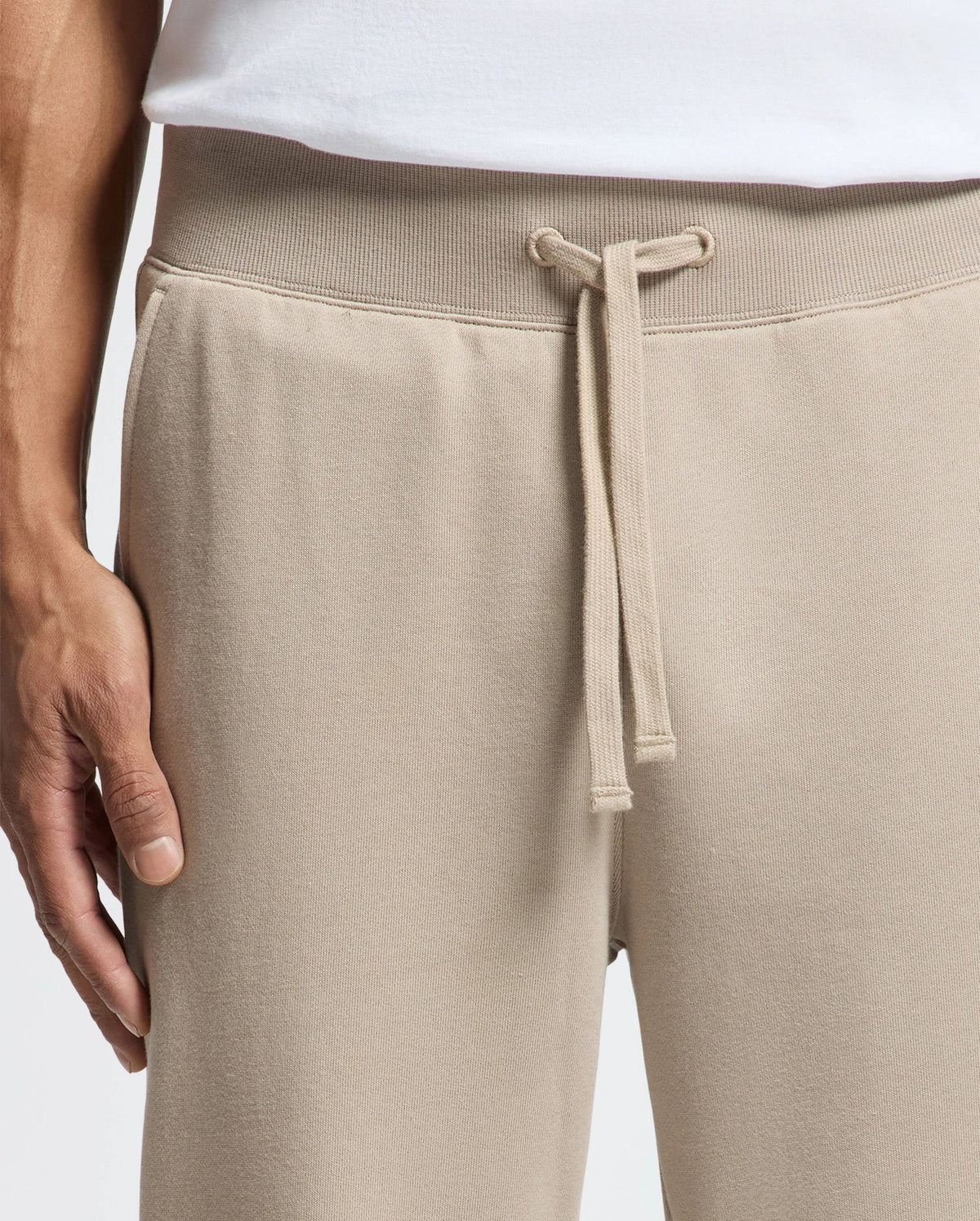 A close-up of a person's hand resting on cozy Psycho Bunny MENS LEVY ESSENTIAL FLEECE SWEATPANT, featuring a drawstring waist, set against a plain background. They are wearing a white shirt.