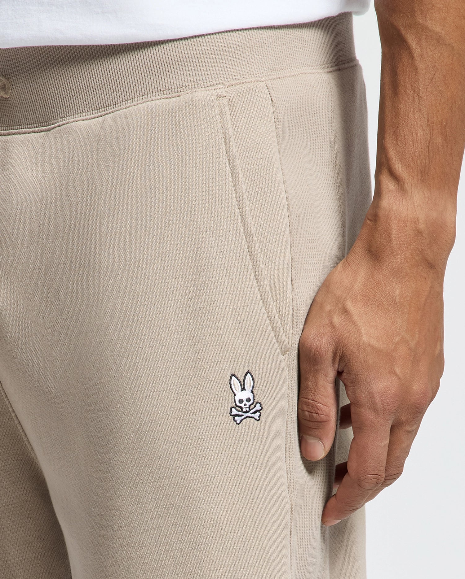 A person is wearing the Psycho Bunny MENS LEVY ESSENTIAL FLEECE SWEATPANT, featuring a discreet embroidered bunny and crossbones logo on the pocket. They are standing with their hand resting near the logo, while a white shirt peeks out from above.