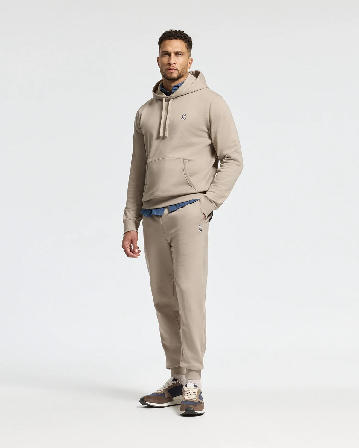 A man is wearing a beige hoodie and matching Psycho Bunny MENS LEVY ESSENTIAL FLEECE SWEATPANT (B6P721D200) joggers, with a blue shirt peeking out underneath. He completes the look with brown and beige sneakers against a plain white background.