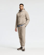 A man is wearing a beige hoodie and matching Psycho Bunny MENS LEVY ESSENTIAL FLEECE SWEATPANT (B6P721D200) joggers, with a blue shirt peeking out underneath. He completes the look with brown and beige sneakers against a plain white background.