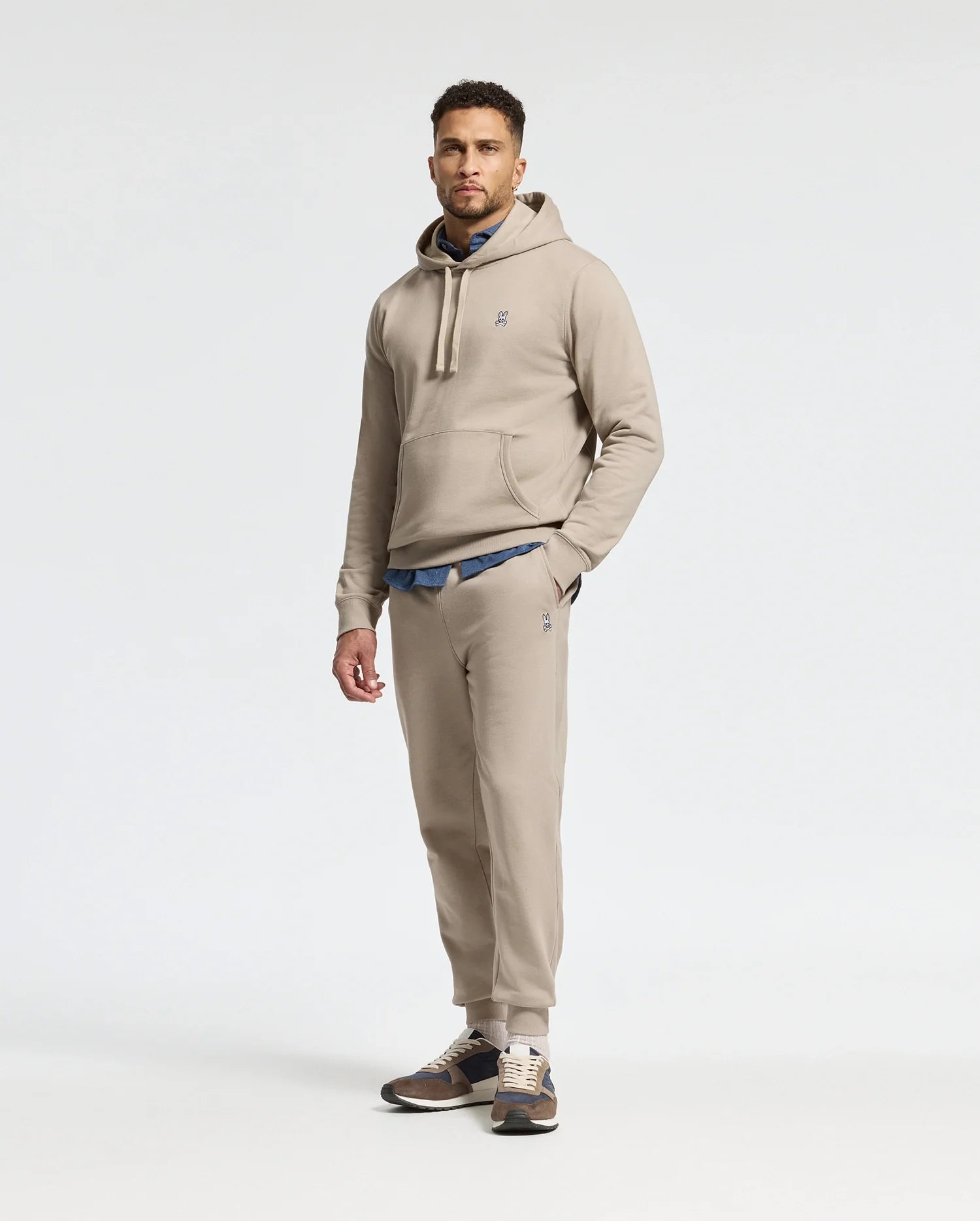 A man is wearing a beige hoodie and matching Psycho Bunny MENS LEVY ESSENTIAL FLEECE SWEATPANT (B6P721D200) joggers, with a blue shirt peeking out underneath. He completes the look with brown and beige sneakers against a plain white background.
