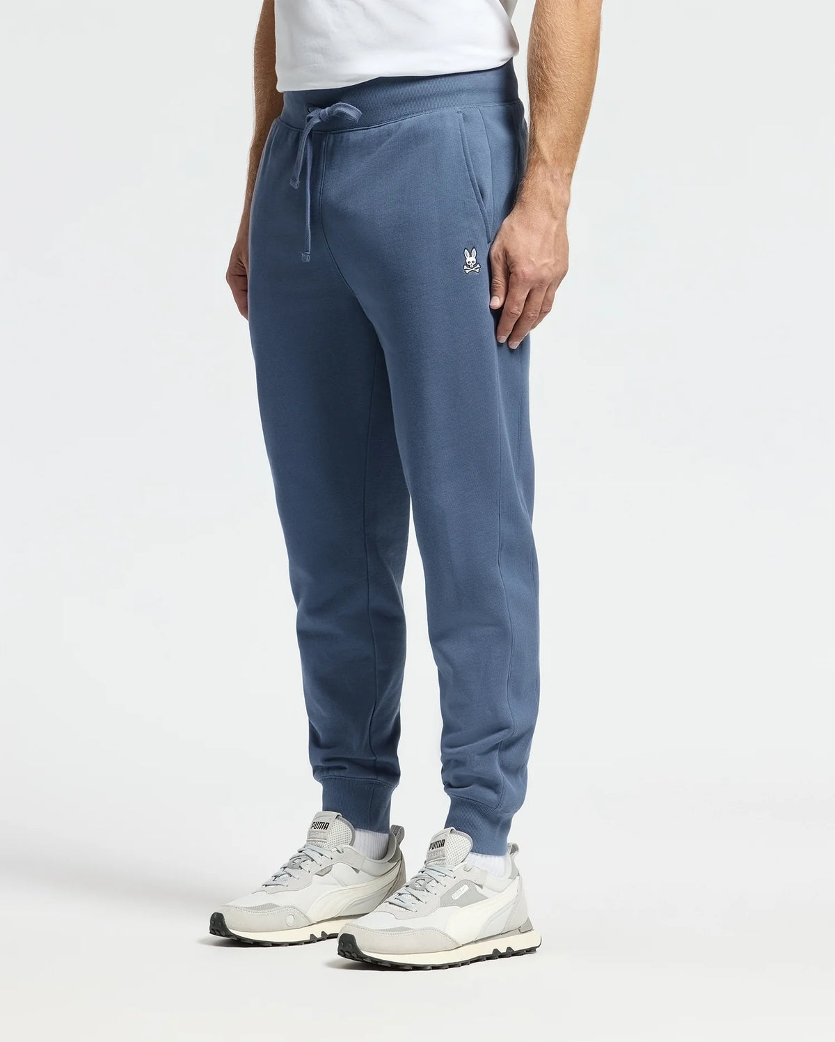 A person is wearing Psycho Bunny MENS LEVY ESSENTIAL FLEECE SWEATPANT - B6P721D200 in blue, paired with white sneakers that have gray accents, and a white t-shirt while standing against a plain background. The sweatpants feature an embroidered logo on the left thigh.