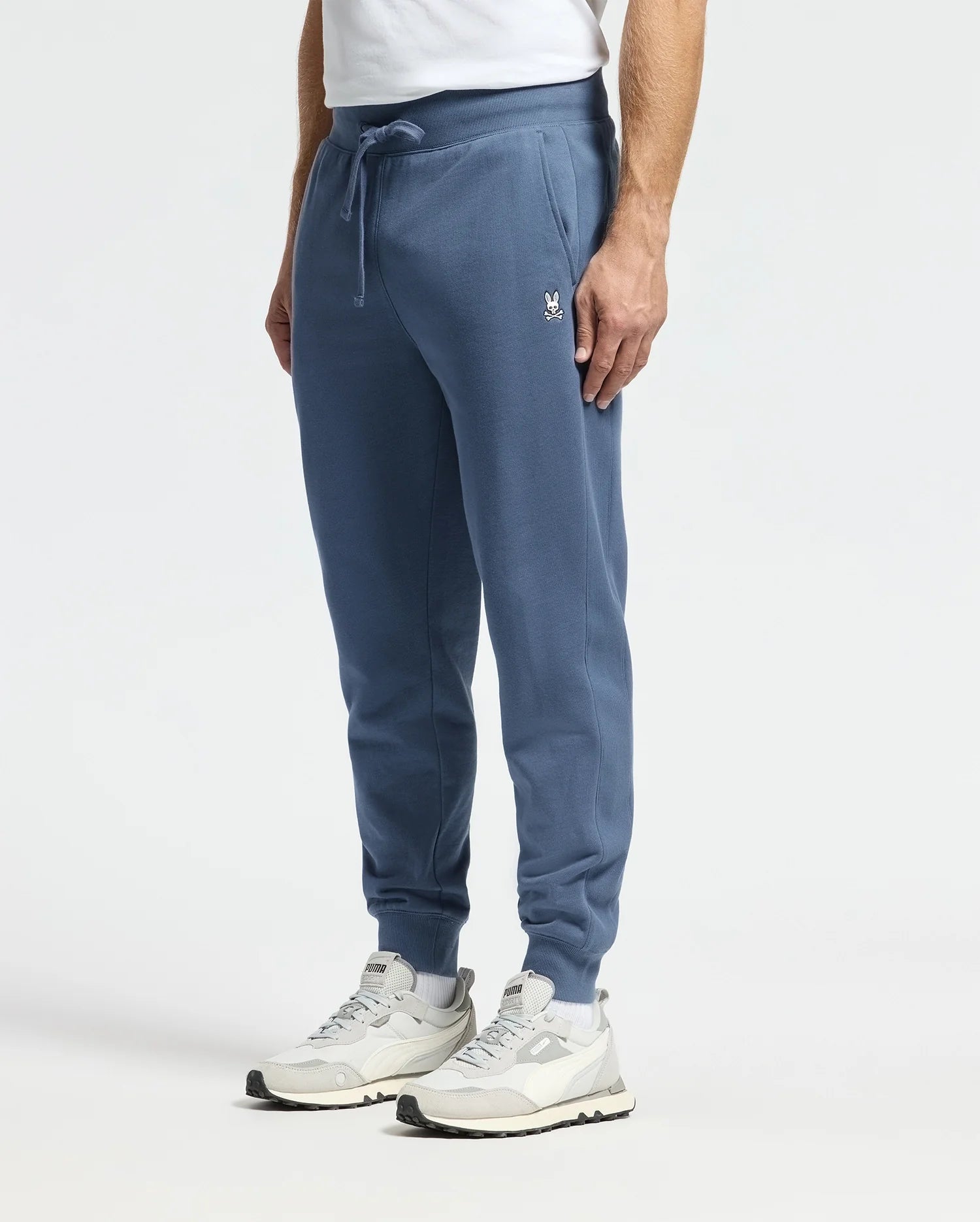 A person is wearing Psycho Bunny MENS LEVY ESSENTIAL FLEECE SWEATPANT - B6P721D200 in blue, paired with white sneakers that have gray accents, and a white t-shirt while standing against a plain background. The sweatpants feature an embroidered logo on the left thigh.