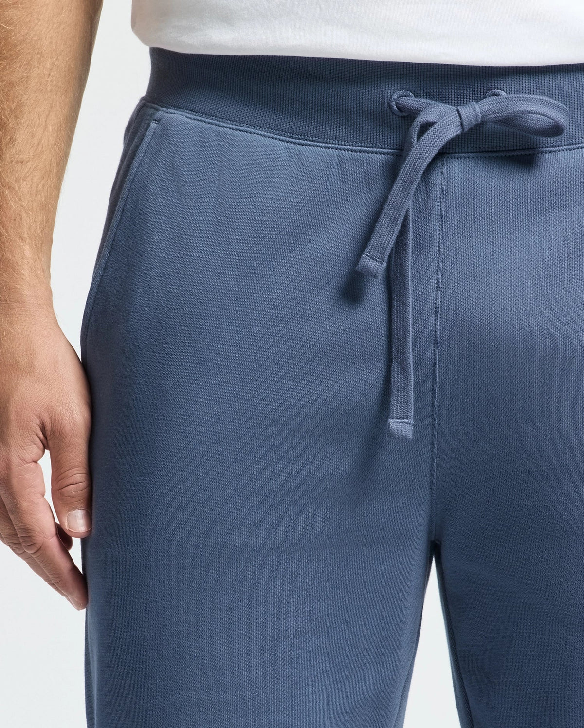 Close-up of a person wearing Psycho Bunny's MENS LEVY ESSENTIAL FLEECE SWEATPANT, featuring an adjustable drawstring. The person is also wearing a white shirt, with their left hand visible resting at the side.