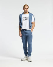 A person stands wearing a white T-shirt with graphic text and comfortable Psycho Bunny MENS LEVY ESSENTIAL FLEECE SWEATPANTS. A matching blue sweatshirt is draped over their shoulders, paired with white sneakers. The background is plain white.