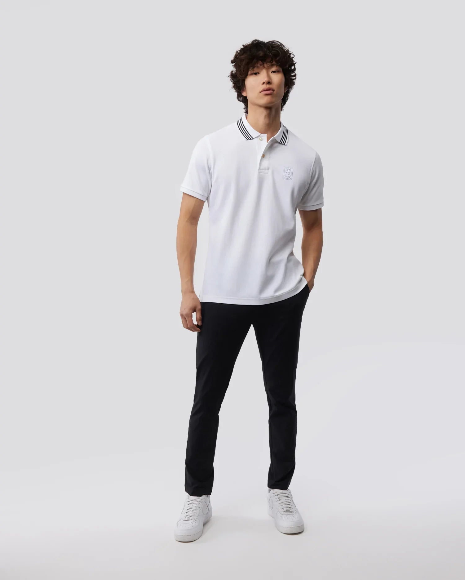A young man in a white polo shirt and Psycho Bunny's MEN'S GABLE REGULAR FIT SPORT PANT - B6P760ARCN stands against a plain light background, looking slightly to the side. His casual, poised appearance is completed with white sneakers.