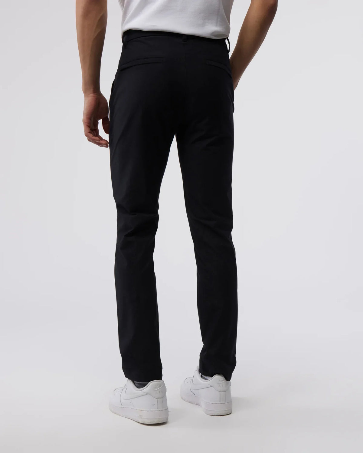 A man stands facing away from the camera, showing the back view of Psycho Bunny's Men's Gable regular fit sport pants paired with white sneakers on a light neutral background.