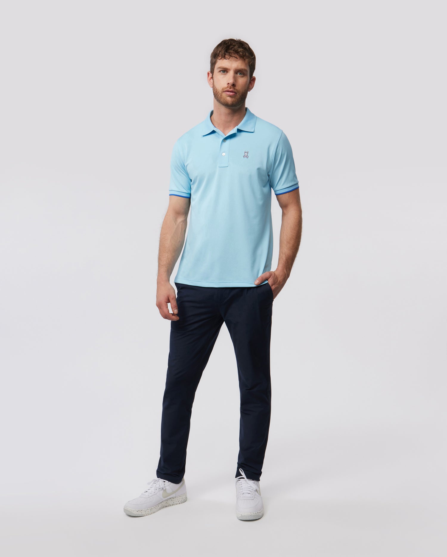 A man stands against a plain background, dressed in a light blue polo shirt, Psycho Bunny's Men's Gable Regular Fit Sport Pants, and white sneakers, looking directly at the camera.