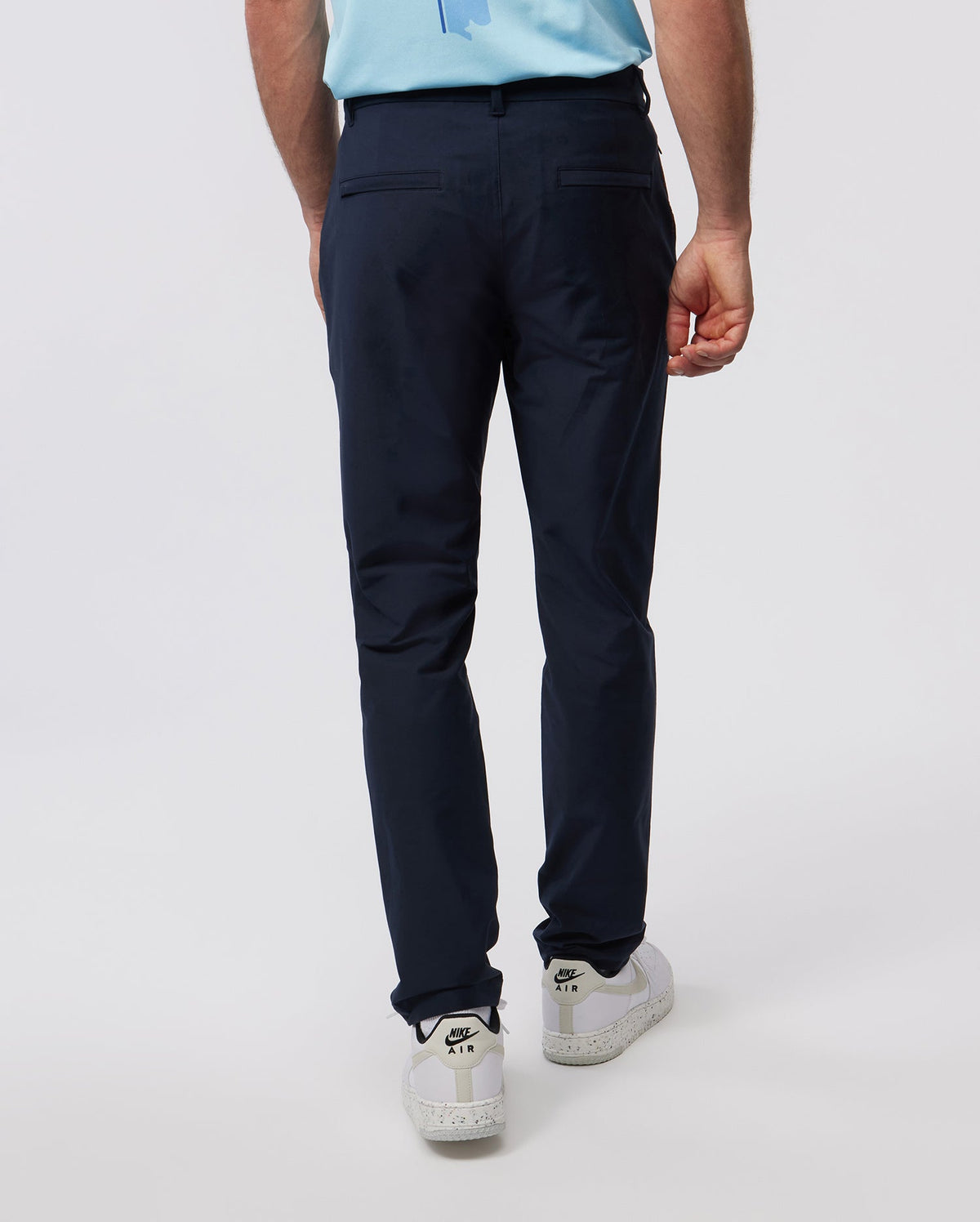 A man from the back wearing Psycho Bunny Men's Gable Regular Fit Sport Pants in navy blue stretch cotton-nylon textile and white sneakers on a plain background, focusing on his lower half.