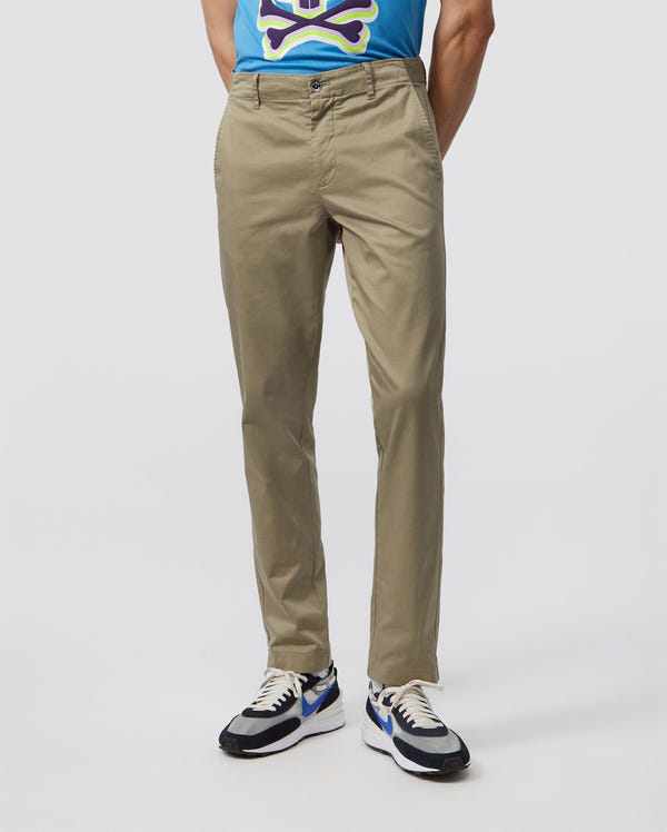 A person wearing Psycho Bunny's MENS DELANCY CHINO PANTS in classic-fit stretch cotton, teamed with a blue shirt with a colorful graphic, and black and white sneakers. The focus is on the lower half of the outfit against a plain background.