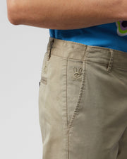 A close-up shows a person wearing Psycho Bunny's classic-fit MENS DELANCY CHINO PANTS in light brown stretch cotton, featuring embroidered rabbit logos near the pocket, paired with a blue shirt against a plain white background.