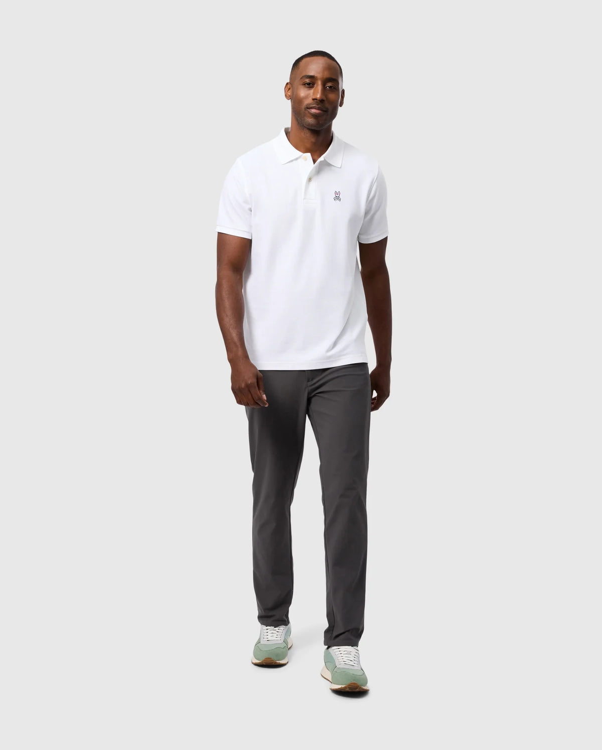 A man stands against a plain light gray background, wearing a white polo shirt and dark gray Psycho Bunny MENS 10K STEP PANT - B6P788D200. He is also wearing light green sneakers with white soles. The pants, featuring a convenient five-pocket design and four-way stretch fabric, complement his neutral expression as he looks forward.