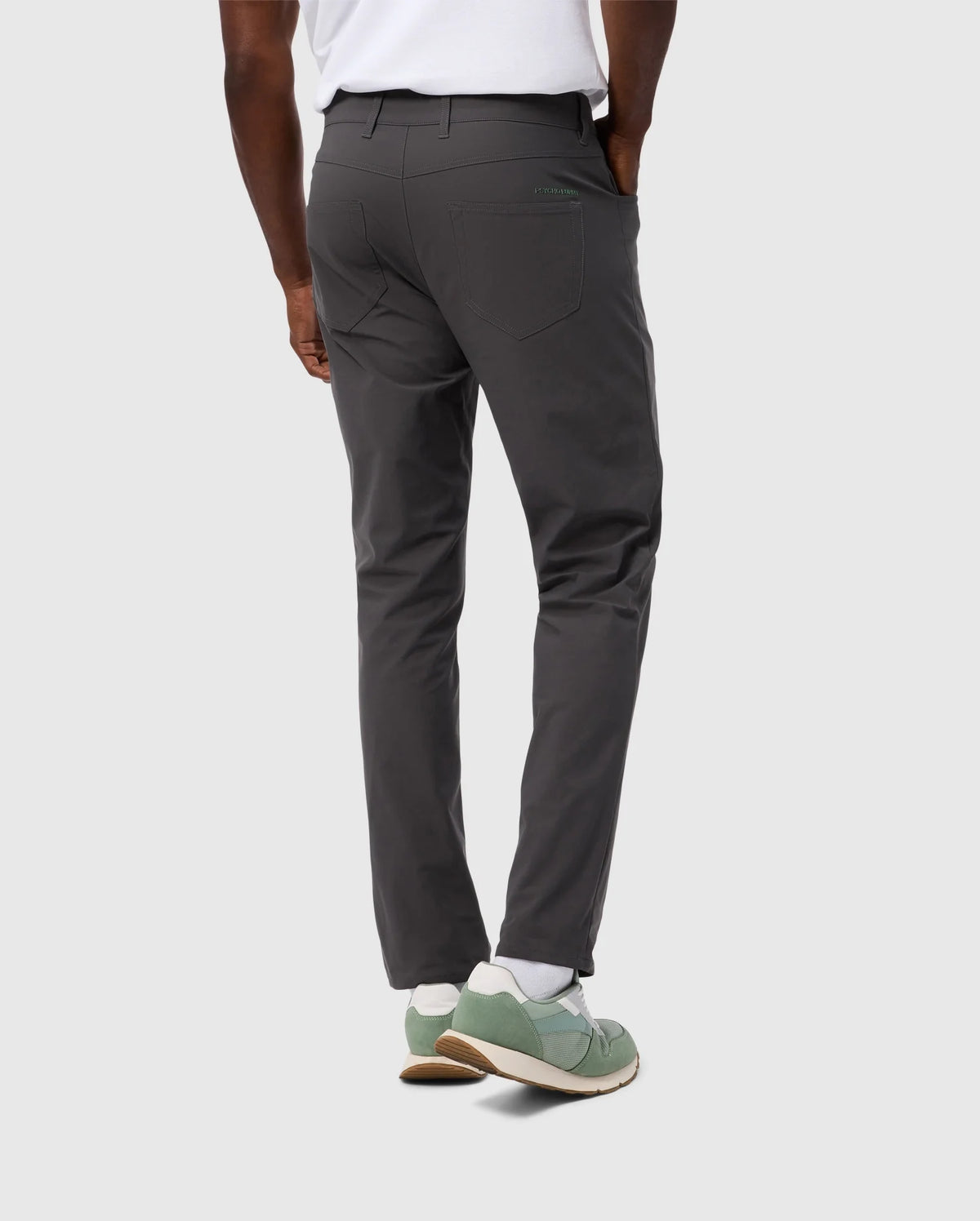 A person stands facing away, clad in a white shirt and Psycho Bunny MEN'S 10K STEP PANT - B6P788D200 in gray, showcasing their four-way stretch. Their right hand rests casually in one of the five pockets, set against a plain gray background, accentuated by green sneakers.