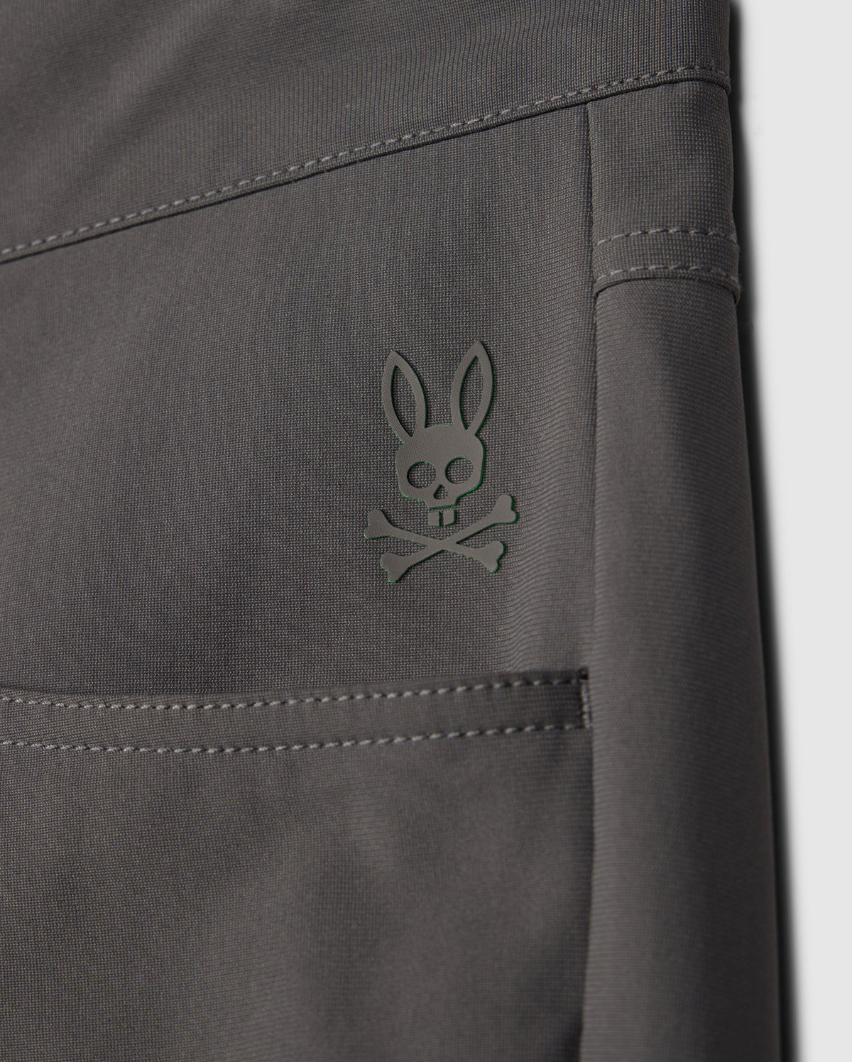 Close-up of the dark gray fabric of the MENS 10K STEP PANT - B6P788D200 by Psycho Bunny, displaying a small embroidered logo. The logo features a stylized bunny head with crossed bones underneath, reminiscent of a skull and crossbones design. The stitching and texture of the four-way stretch fabric are clearly visible.