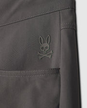 Close-up of the dark gray fabric of the MENS 10K STEP PANT - B6P788D200 by Psycho Bunny, displaying a small embroidered logo. The logo features a stylized bunny head with crossed bones underneath, reminiscent of a skull and crossbones design. The stitching and texture of the four-way stretch fabric are clearly visible.