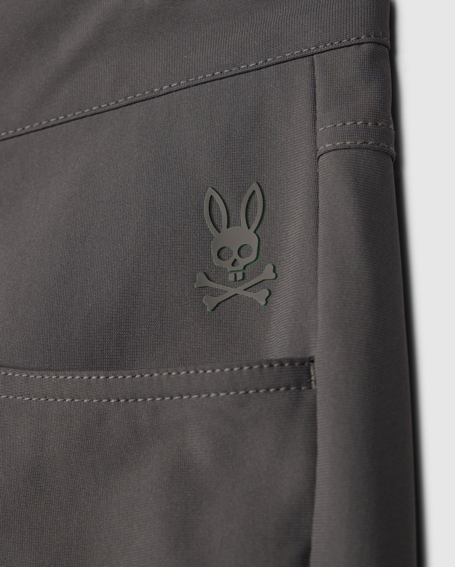 Close-up of the dark gray fabric of the MENS 10K STEP PANT - B6P788D200 by Psycho Bunny, displaying a small embroidered logo. The logo features a stylized bunny head with crossed bones underneath, reminiscent of a skull and crossbones design. The stitching and texture of the four-way stretch fabric are clearly visible.
