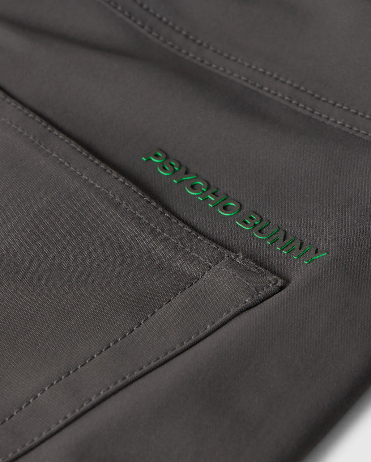 Close-up image of dark gray fabric with visible stitching and the words "Psycho Bunny" embroidered in green thread, showcasing the exceptional quality of our MENS 10K STEP PANT - B6P788D200's four-way stretch material.