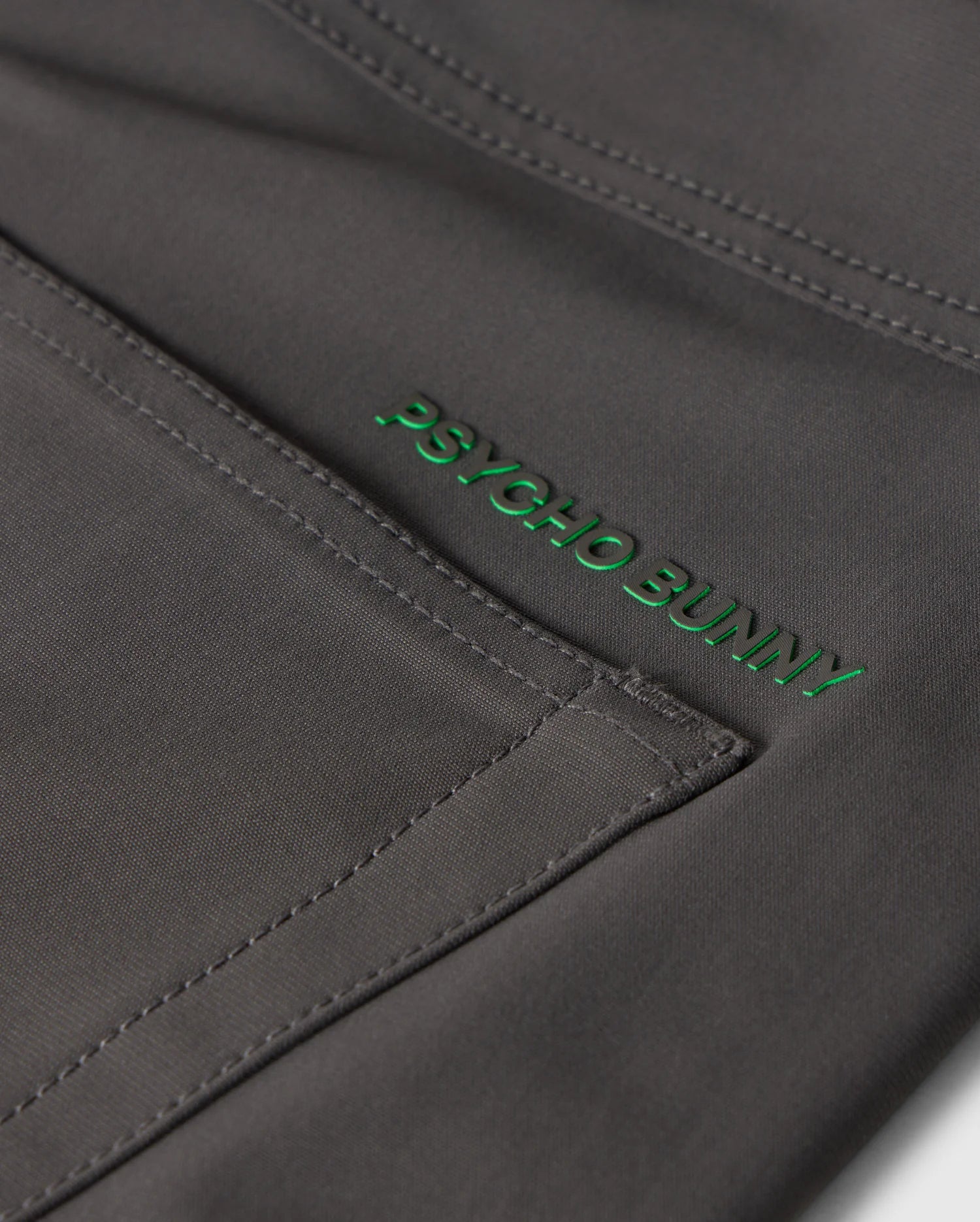Close-up image of dark gray fabric with visible stitching and the words "Psycho Bunny" embroidered in green thread, showcasing the exceptional quality of our MENS 10K STEP PANT - B6P788D200's four-way stretch material.