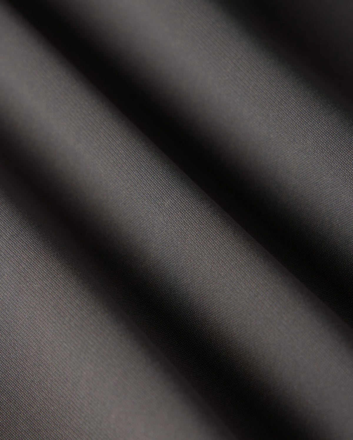 Close-up of smooth, dark grey fabric with soft, diagonal folds creating light and shadow contrast. The texture appears fine and slightly reflective, reminiscent of the luxurious material used in Psycho Bunny's versatile MENS 10K STEP PANT - B6P788D200.