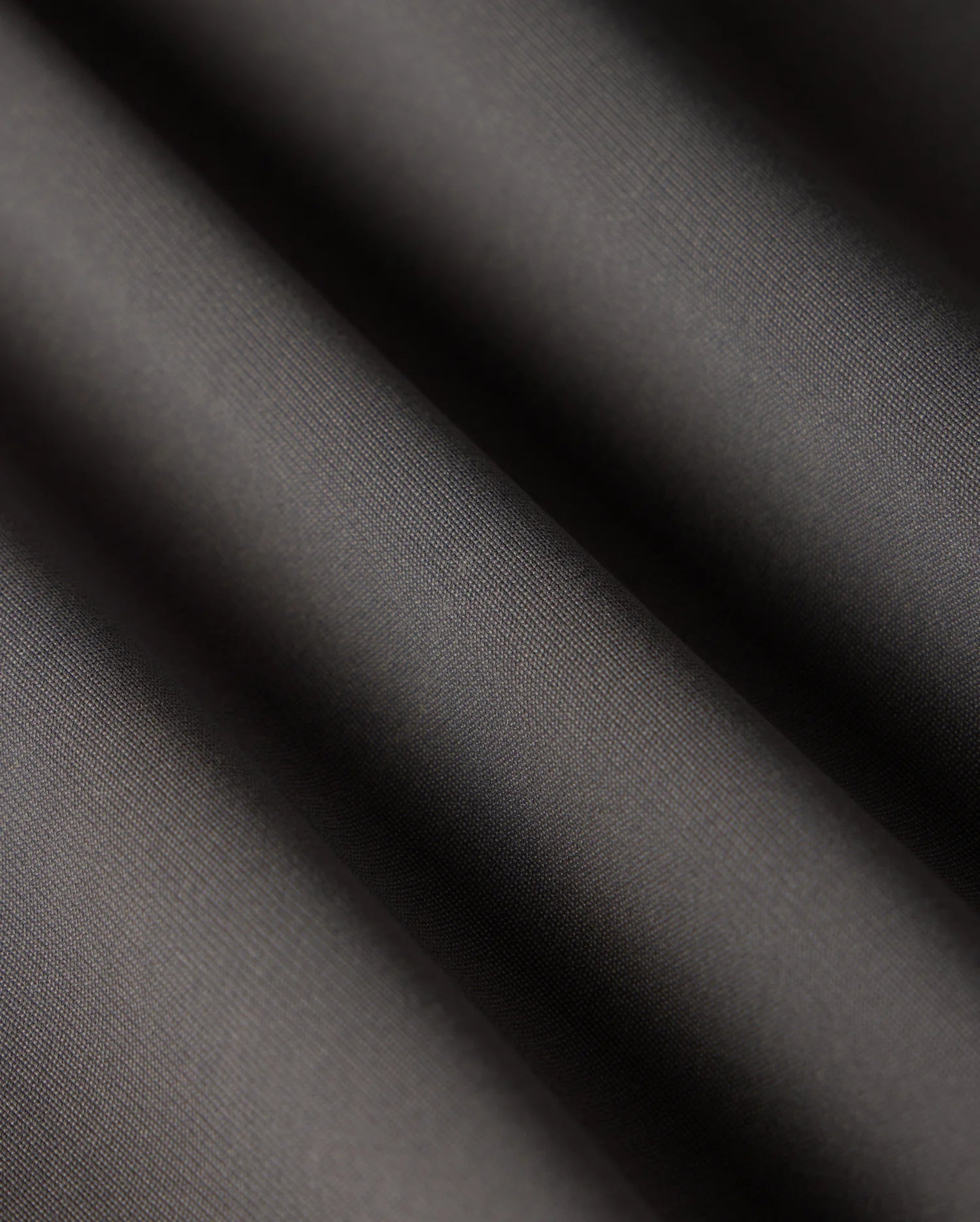 Close-up of smooth, dark grey fabric with soft, diagonal folds creating light and shadow contrast. The texture appears fine and slightly reflective, reminiscent of the luxurious material used in Psycho Bunny's versatile MENS 10K STEP PANT - B6P788D200.