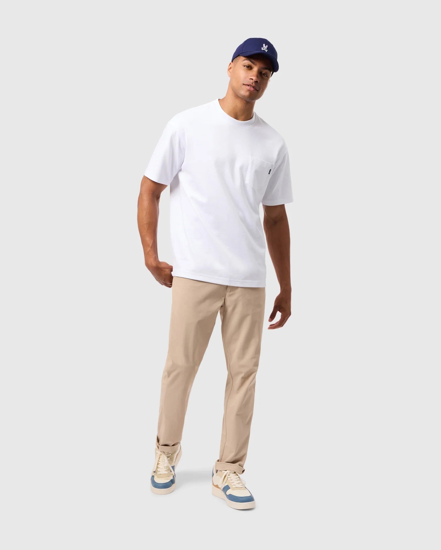 A person is wearing a white T-shirt, the Psycho Bunny MENS 10K STEP PANT - B6P788D200 in beige, a navy blue cap, and light-colored sneakers. They are standing against a plain gray background with a relaxed posture, tilting slightly to one side.