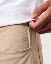 Up-close shot of an individual donning beige Psycho Bunny MENS 10K STEP PANT - B6P788D200, showcasing the five-pocket design and the embroidered bunny skull and crossbones logo. The person slightly lifts a white shirt, revealing the logo on the pants' hip area near a pocket.