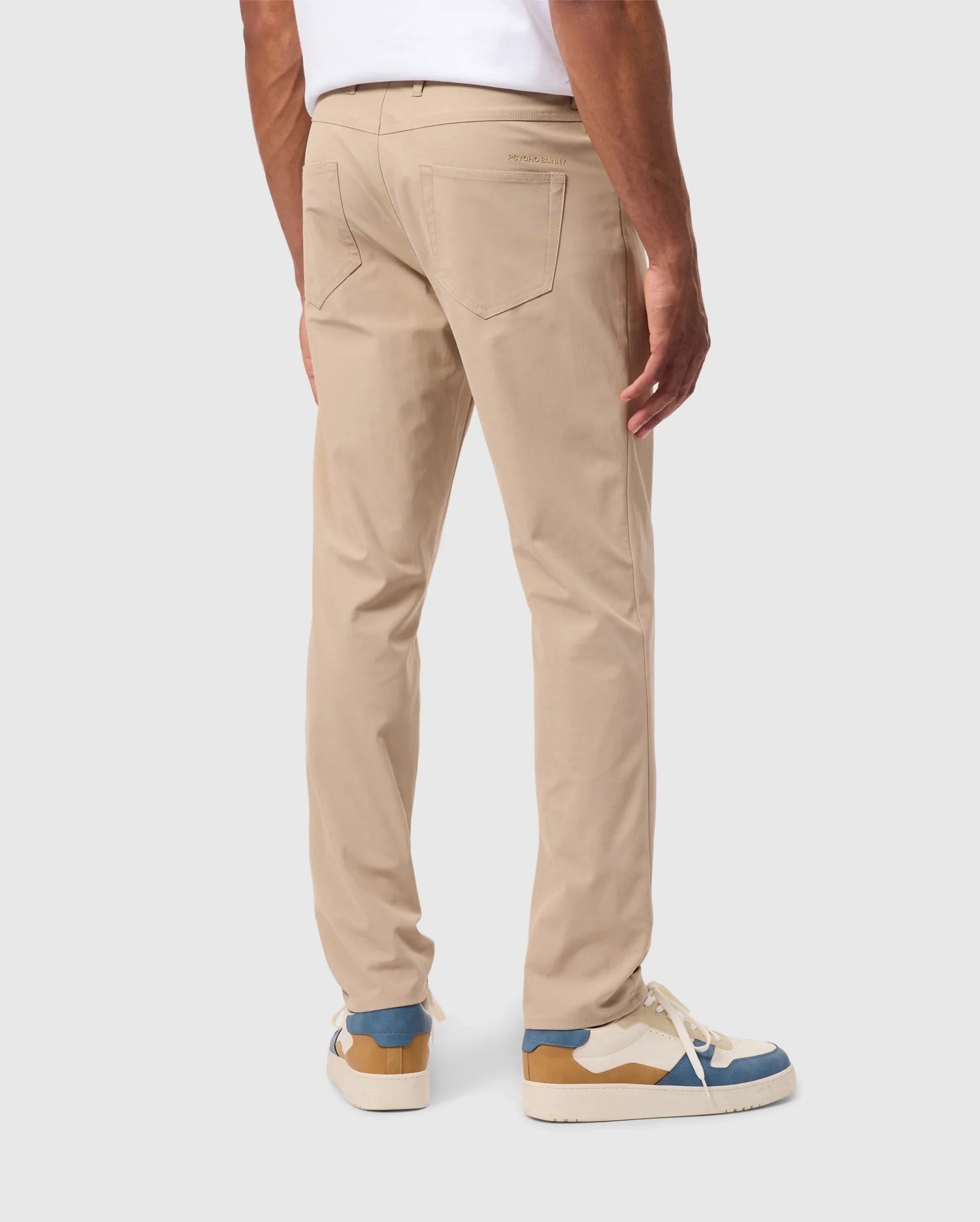 A person is standing, facing away from the camera, dressed in Psycho Bunny's beige MENS 10K STEP PANT (B6P788D200) with a five-pocket design and a white shirt. Their footwear consists of white sneakers with blue and brown accents. The background is plain and light-colored.