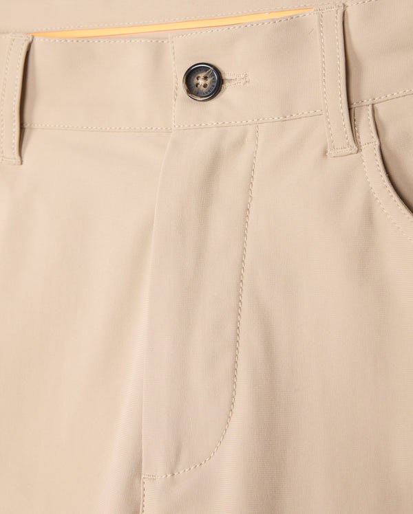 A close-up view of the Psycho Bunny MENS 10K STEP PANT - B6P788D200 beige four-way stretch trousers focusing on the waistband, a black button, and belt loops. The fabric appears smooth and well-stitched, showcasing the front pocket detail and a part of the yellow lining inside the waistband.