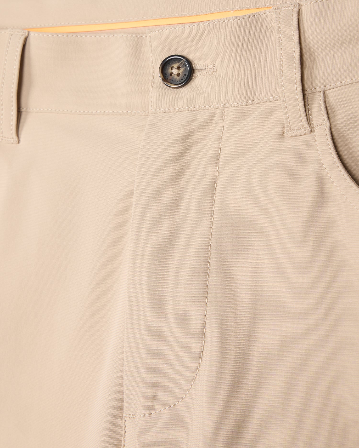A close-up view of the Psycho Bunny MENS 10K STEP PANT - B6P788D200 beige four-way stretch trousers focusing on the waistband, a black button, and belt loops. The fabric appears smooth and well-stitched, showcasing the front pocket detail and a part of the yellow lining inside the waistband.