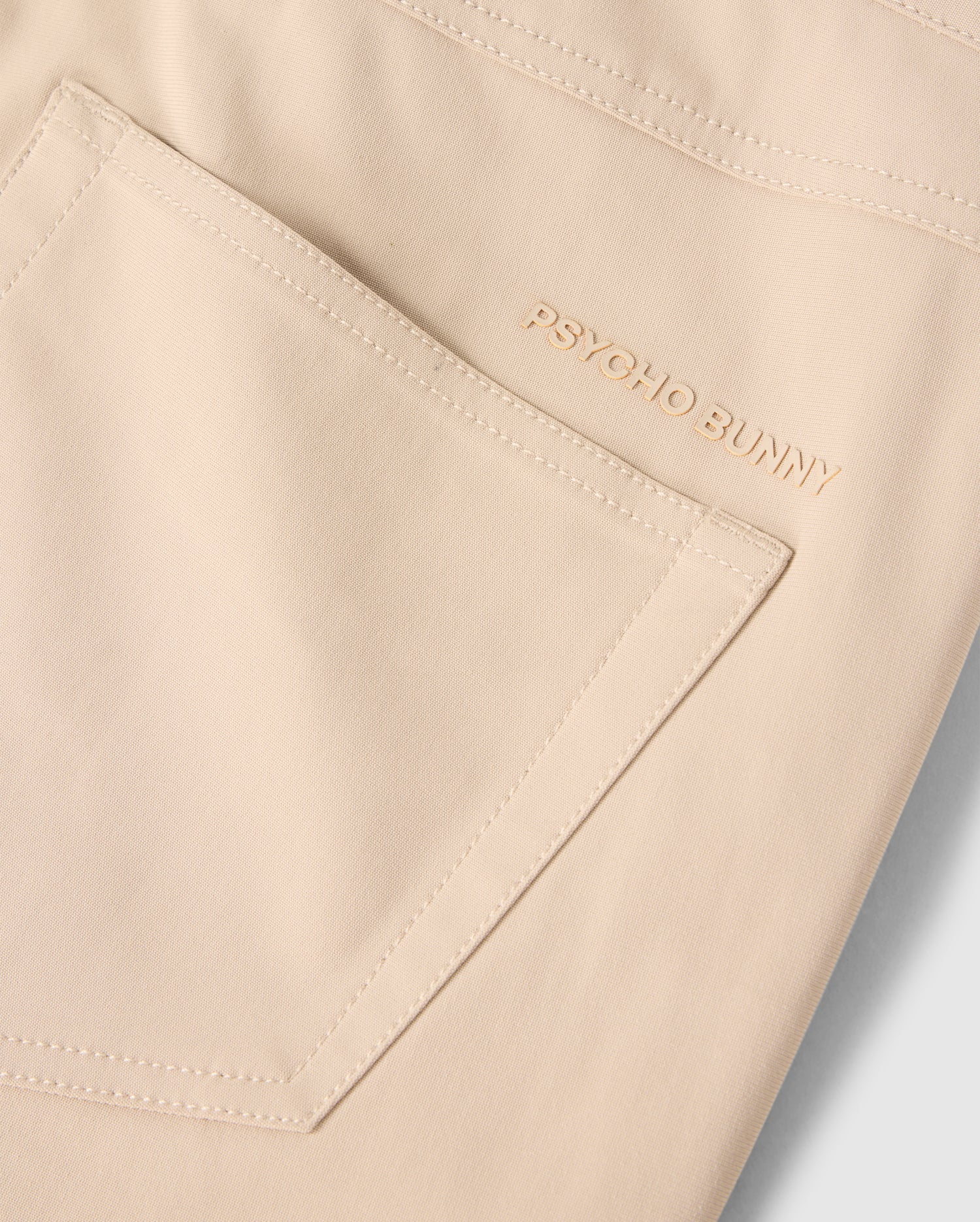 Close-up of the back pocket of a light beige, smooth and slightly glossy MENS 10K STEP PANT - B6P788D200 with the brand name "Psycho Bunny" embroidered in matching beige thread above the pocket. This sleek design features a comfortable four-way stretch for ease of movement.