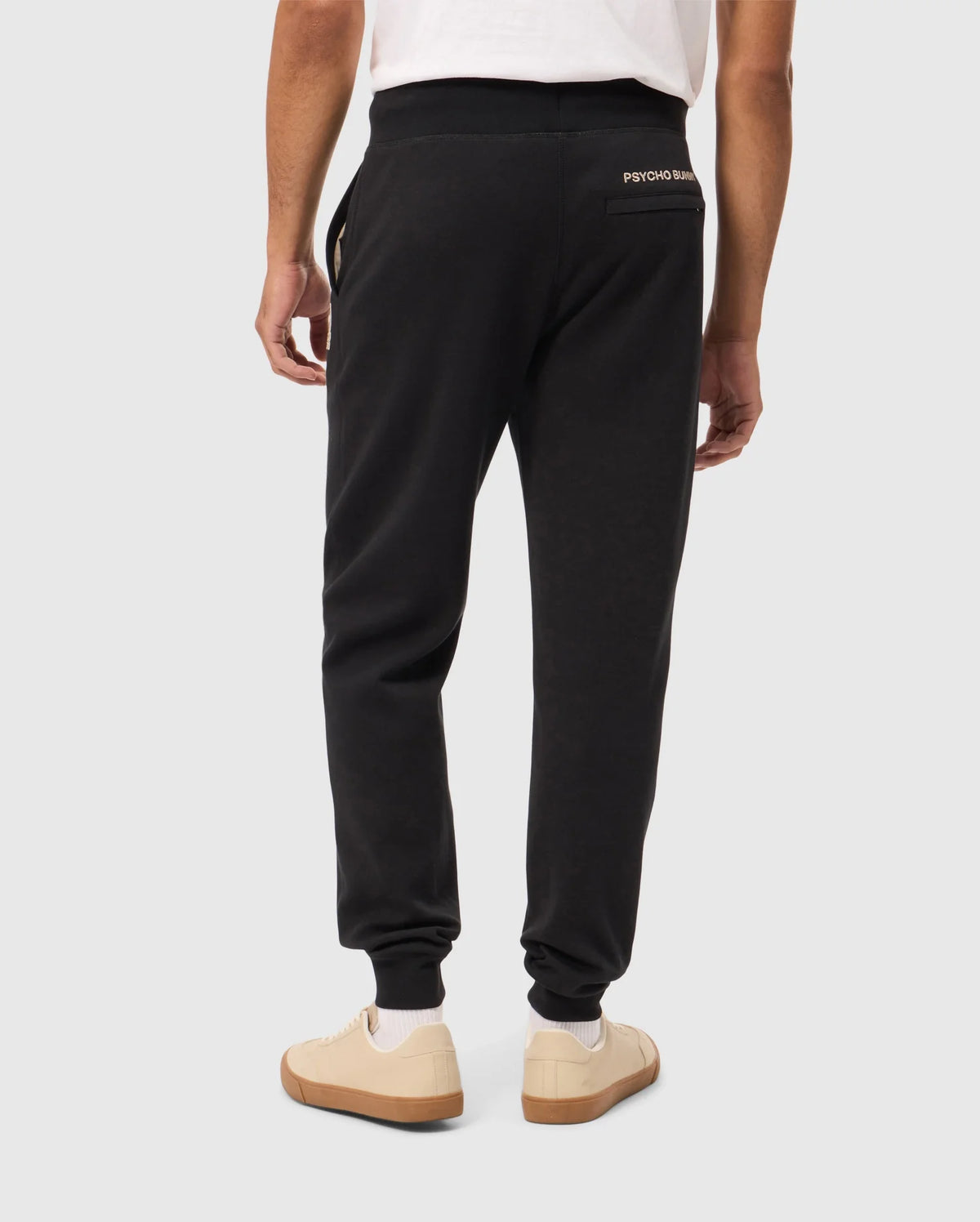 Rear view of a person wearing Psycho Bunny's James Premium Sweatpant in black cotton-blend and a white t-shirt. They stand against a plain background, showcasing white sneakers with tan soles. These premium sweatpants feature the distinctive small logo on the back, near the waistband.