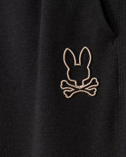 A close-up of the black cotton-blend fabric on the Psycho Bunny MENS JAMES PREMIUM SWEATPANT - B6P791D200 reveals a stitched emblem featuring a bunny head silhouette above crossed bones. This emblem, in light beige, provides a striking contrast that enhances the design. The detailed and precise stitching showcases the craftsmanship of these premium sweatpants.