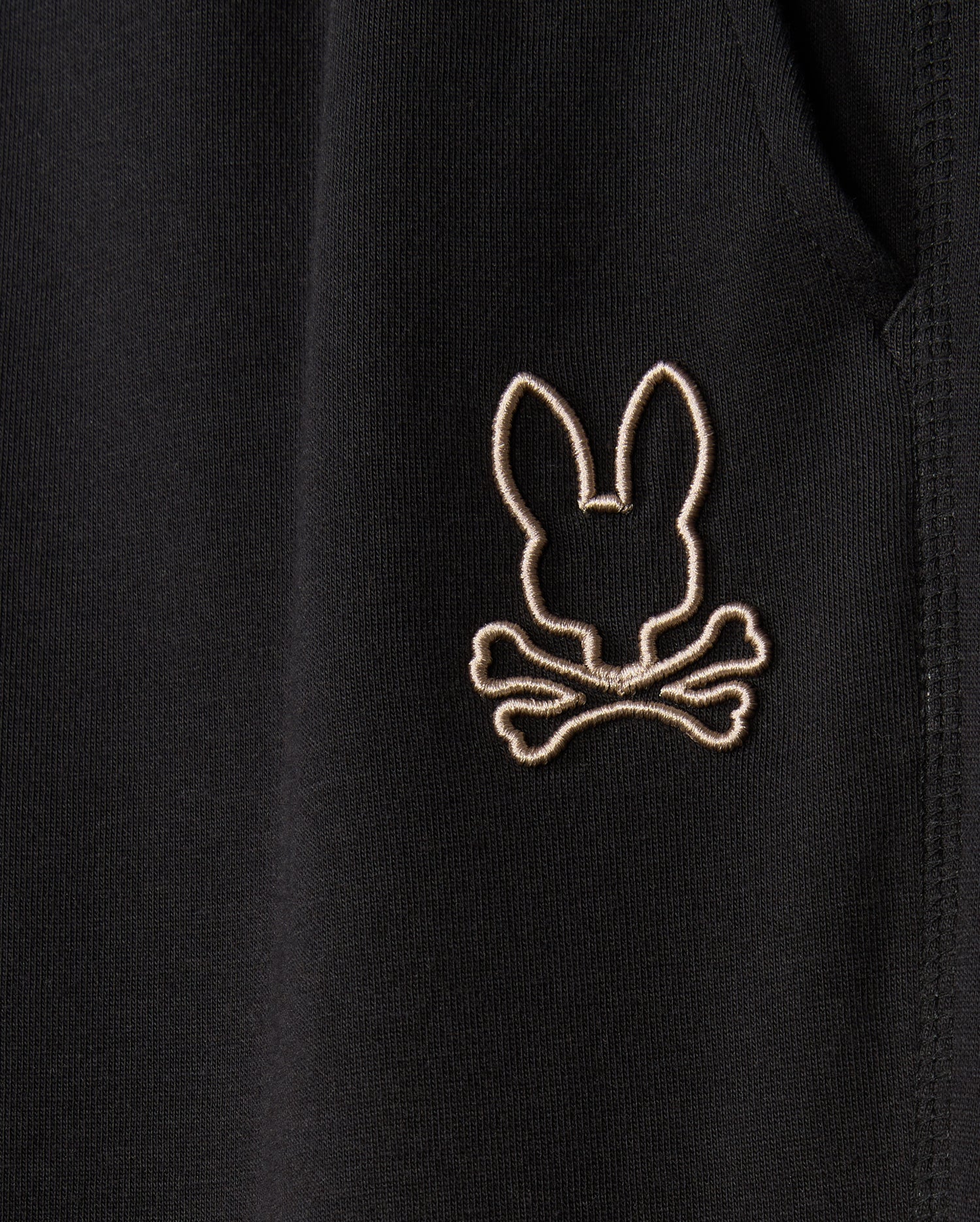 A close-up of the black cotton-blend fabric on the Psycho Bunny MENS JAMES PREMIUM SWEATPANT - B6P791D200 reveals a stitched emblem featuring a bunny head silhouette above crossed bones. This emblem, in light beige, provides a striking contrast that enhances the design. The detailed and precise stitching showcases the craftsmanship of these premium sweatpants.