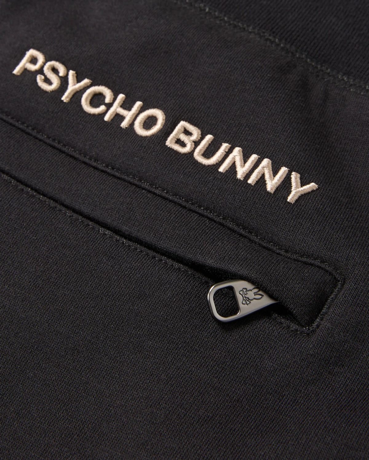 Close-up of a black cotton-blend garment displaying the embroidered text "Psycho Bunny" in white. Below the text, there's a partially open small zipper pocket with a branded logo on the zipper pull, ideal for pairing with premium bottoms such as the MENS JAMES PREMIUM SWEATPANT - B6P791D200.