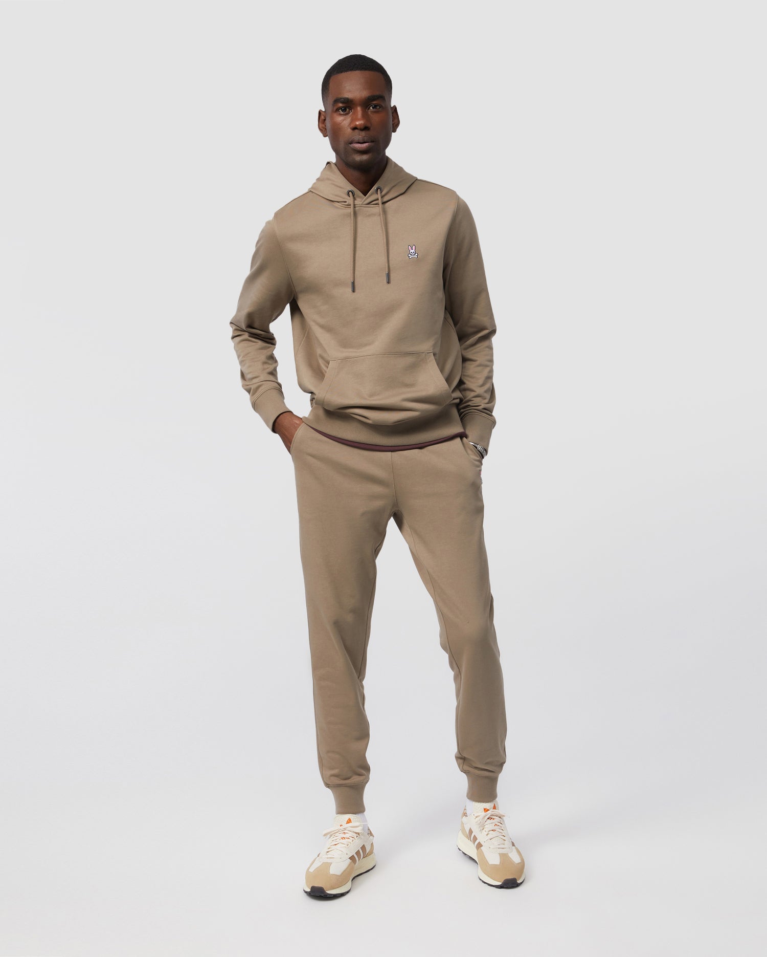 Dressed in Psycho Bunny's MENS FRENCH TERRY KNIT SWEATPANTS, a man stands against a white backdrop. His beige hoodie and matching sneakers complement his short hair, embodying effortless casual style with hands relaxed in pockets.