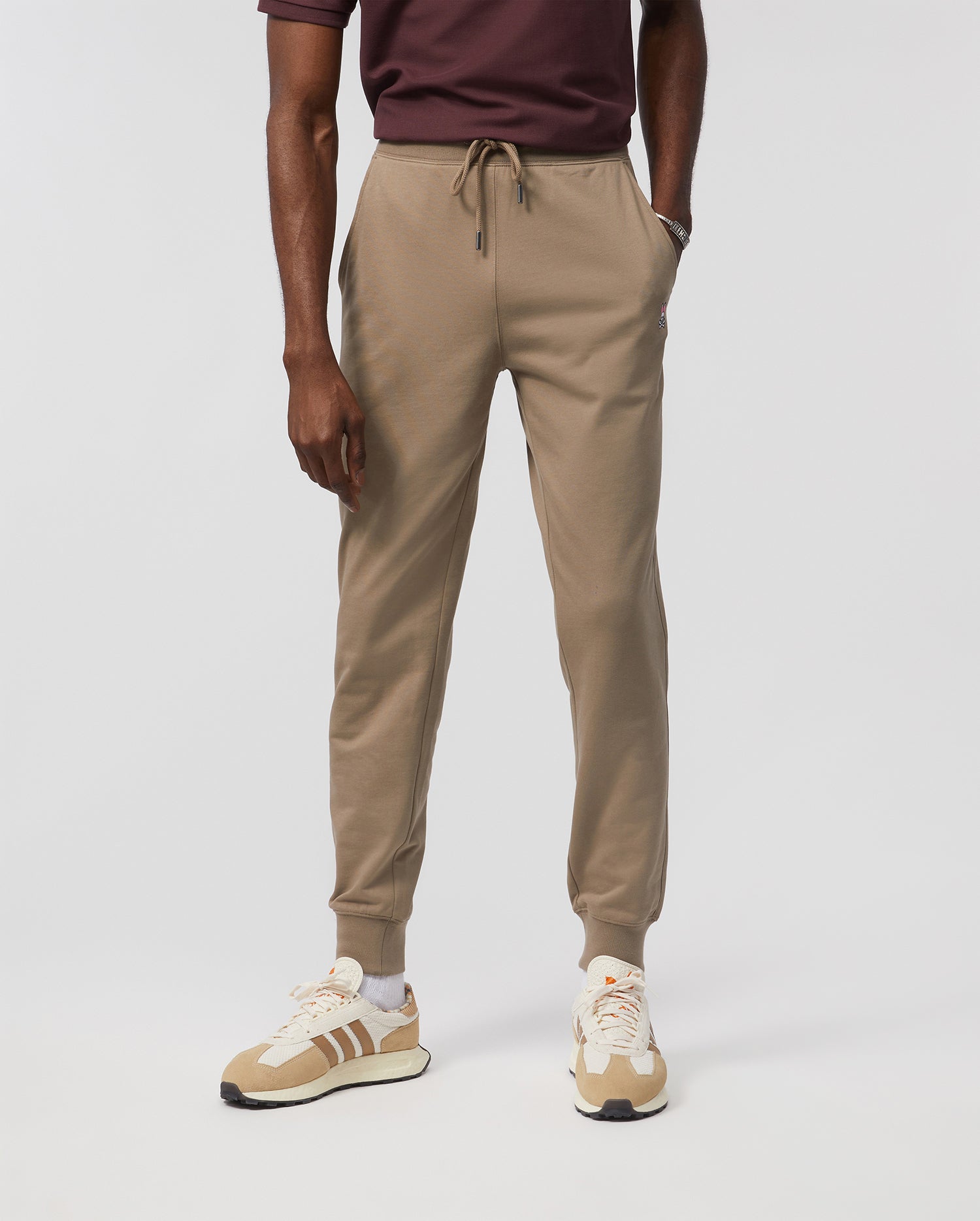 A person is dressed in Psycho Bunny's MENS FRENCH TERRY KNIT SWEATPANTS - B6P828ARFT, which are classic-fit beige joggers. Paired with a maroon shirt and beige sneakers with white laces, the outfit looks casual and sporty against a plain white background.