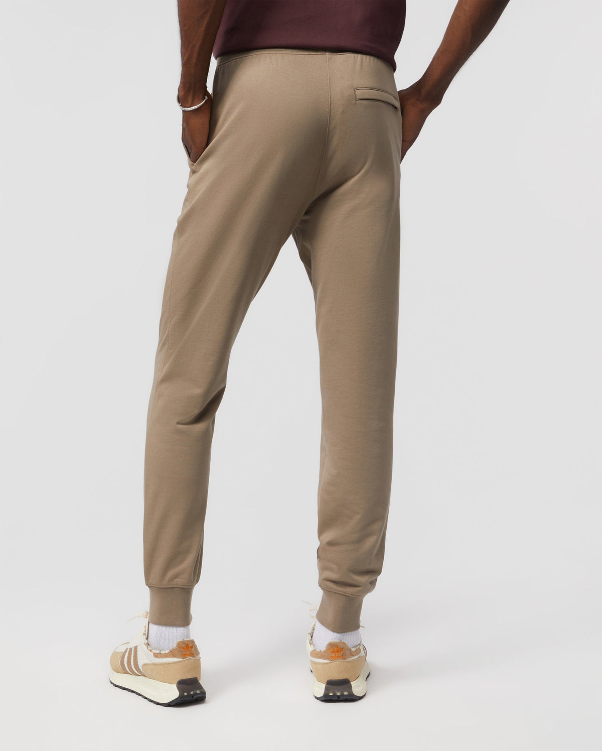 A person wearing Psycho Bunny's men's French terry knit sweatpants (B6P828ARFT) and white sneakers with beige accents stands with their hands in their pockets, showcasing the relaxed fit and back pocket of the joggers, set against a plain white background.