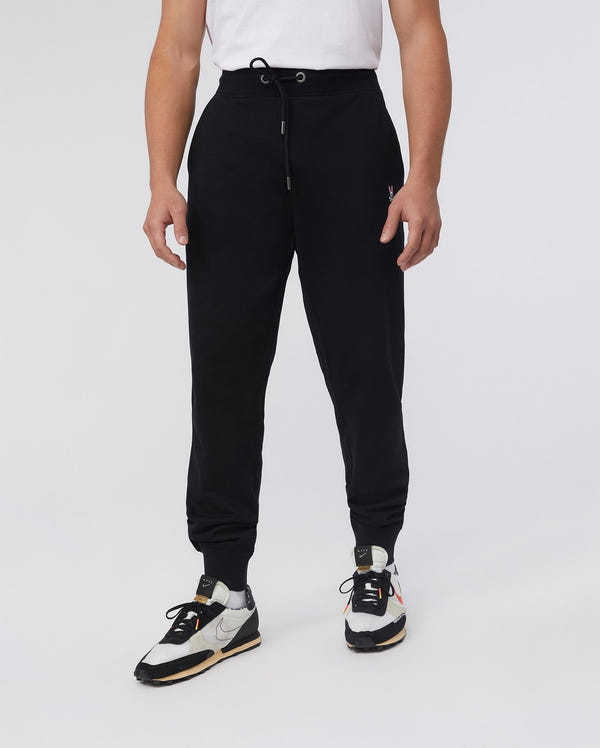 A person stands against a plain background wearing a white t-shirt, classic fit black Psycho Bunny MENS FRENCH TERRY KNIT SWEATPANTS - B6P828ARFT with drawstrings, and black and white sneakers featuring yellow accents.